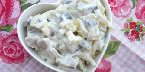 Pyszny przepis na śledzie w śmietanie z jabłkiem i ogórkiem. Sałatka w białej miseczce w kształcie serca na tle różowych róż.