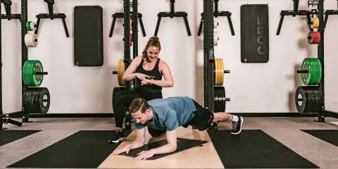 Ein Mann macht einen Unterarmstütz, während eine Trainerin die Zeit stoppt. Perfekt für einen HIIT Trainingsplan im Fitnessstudio.