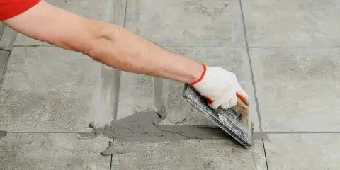 Mano con guante aplica lechada gris a las juntas entre azulejos de cemento con una llana.