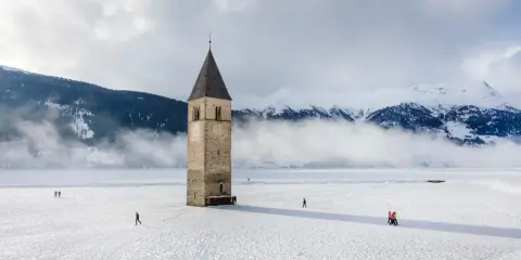 Torre sommersa nel lago di Resia, circondata da neve e montagne nebbiose. Alcune persone passeggiano sul ghiaccio.