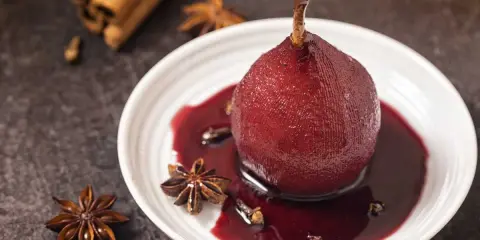 Peras al vino tinto, cocidas con especias como anís estrellado y canela, servidas en un plato blanco con su sirope.