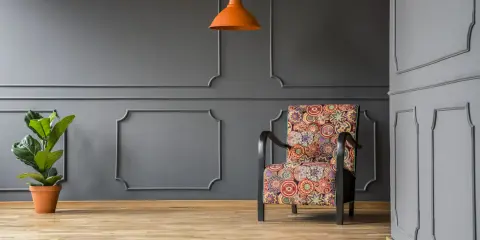 Nowoczesne sztukateria ścienna inspiracje: fotel w kolorowe wzory, pomarańczowa lampa i roślina na tle szarej ściany.