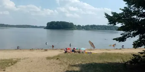 Słoneczny dzień nad jeziorem przeczyckim. Ludzie odpoczywają na piaszczystej plaży, kąpią się i pływają.
