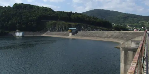 Betonowa zapora spiętrza wody Jeziora Żywieckiego. W oddali zielone wzgórza i błękitne niebo z chmurami.