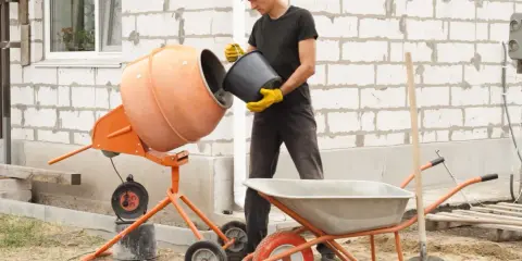 Budowlaniec wsypuje składniki do betoniarki, przygotowując beton B30. W tle taczka i łopata.