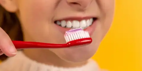 Uśmiechnięta twarz z białymi zębami, na czerwonej szczoteczce pasta do zębów. Dbanie o higienę to podstawa, jak lakierowanie zębów.