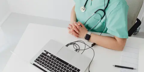Lekarka w zielonym fartuchu z latarką i laptopem przed sobą. Czy do ginekologa potrzebne jest skierowanie? Zapytaj online.