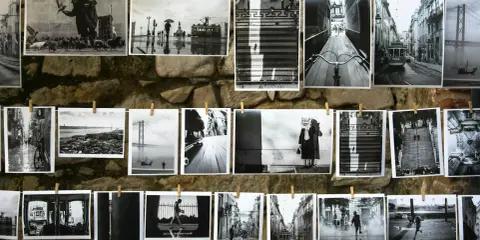 Czarno-białe fotografie wiszą na sznurku na kamiennej ścianie. Widać na nich uliczne sceny z Lizbony, w tym tramwaje, most i ludzi.