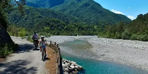 Ciclovia del Tagliamento: due ciclisti esplorano il sentiero panoramico lungo il fiume, circondati da montagne verdi e un cielo azzurro.