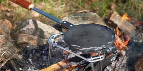 En gjutjärnspanna med trähandtag står på en ställning över en öppen eld. Perfekt för att steka plättar eller våfflor ute i naturen.