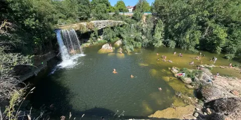 Gente disfrutando de un día soleado en las pozas del río Tus, con una cascada y vegetación exuberante.