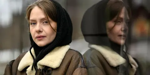 Sofia Kappel i en brun fårskinnsjacka med svart luva, blickar ut. Hennes spegelbild syns i en reflekterande yta.