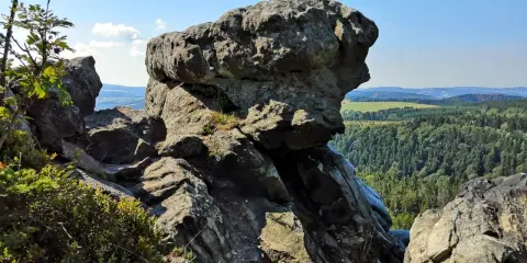 Wielkie, szare skały górują nad zieloną doliną. W oddali widać pola i lasy, a na niebie błękit i chmury.