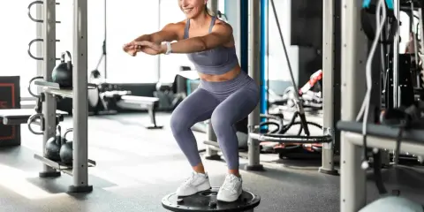 Frau macht Kniebeugen auf einem Bosu-Ball, trägt Sportkleidung und weiße Sneaker. Sie trainiert ohne schuhe auf dem instabilen Untergrund.