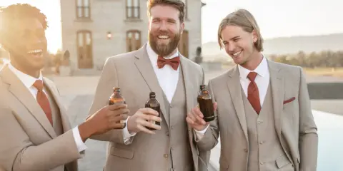 Groomsmen in tan suits with rust ties and bowties toast with beers. A great color suit for wedding celebrations.