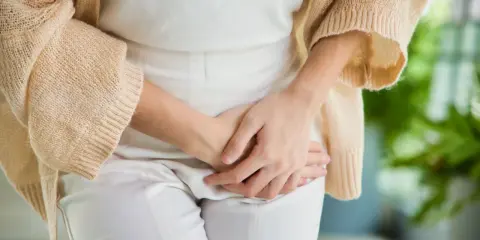 Kobieta w białych spodniach i beżowym swetrze trzyma dłonie na podbrzuszu, odczuwając parcie na mocz.