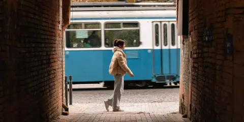 Una ragazza cammina in un vicolo, un tram blu sfreccia sullo sfondo. Una foto vedo non vedo che cattura un momento fugace.