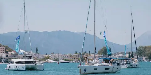 Trois voiliers naviguent dans une baie ensoleillée. Au premier plan, un catamaran monocoque blanc avec des voiles bleues et blanches. Derrière, un autre voilier blanc avec des personnes à bord. Au loin, une ville côtière et des montagnes.