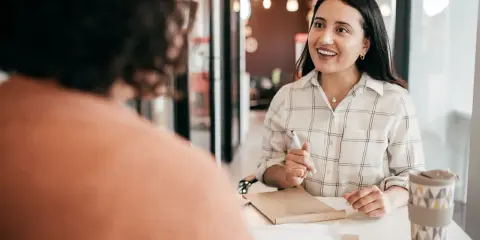 Uśmiechnięta kobieta podczas rozmowy kwalifikacyjnej po angielsku, odpowiada na pytania. Notuje coś w zeszycie.