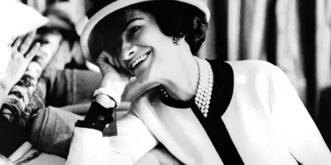 Coco Chanel, sonriendo con un sombrero y perlas, evoca la elegancia de donde es Chanel.