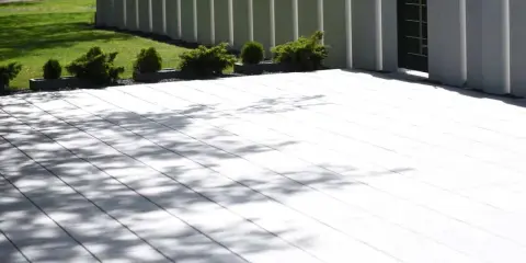 Nowoczesny taras z płyt betonowych, otoczony zielenią i nowoczesnym ogrodzeniem. Cienie drzew tańczą na jasnej nawierzchni.