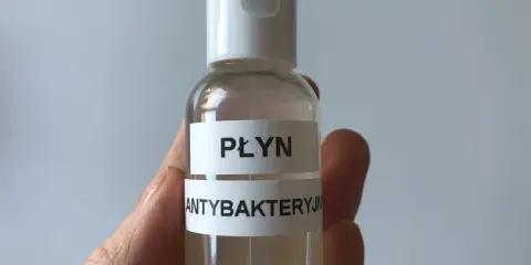 Dłoń trzyma małą, przezroczystą butelkę z białym zamknięciem, zawierającą płyn.