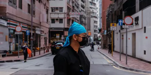 Uomo con bandana blu e mascherina nera in una foto di strada con edifici alti e traffico.