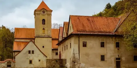 Czerwony klasztor w Pieninach, z charakterystyczną wieżą zegarową i pomarańczowymi dachami, otoczony zielonym trawnikiem i drzewami.