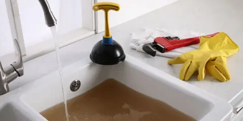 Un évier bouché après siphon, avec de l'eau sale stagnante. Un déboucheur, une clé à molette rouge et des gants jaunes sont prêts à l'emploi.