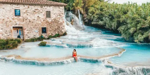 Cascate naturali con acqua turchese, una donna seduta su una roccia. Un antico mulino in pietra sullo sfondo. Tra le terme più belle d'Italia.