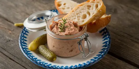 Un pot de rillettes de porc, garni d'une brindille de romarin, est servi avec des cornichons et des tranches de pain croustillant sur une assiette décorée.