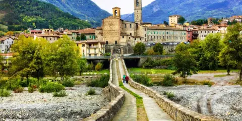 Ponte Gobbo e borghi vicino a Bobbio, con montagne sullo sfondo. Due persone passeggiano sul ponte antico.