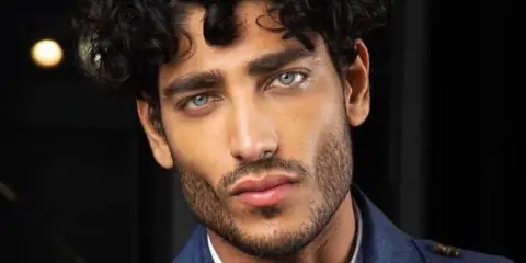 Primo piano di un uomo con splendidi capelli ricci scuri e occhi azzurri penetranti, indossa una giacca blu.