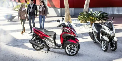 Dos motocicletas Yamaha Tricity, una roja y otra blanca, listas para circular. Tres personas pasean al fondo.