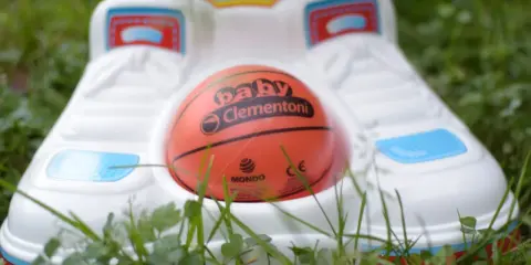 Pomarańczowa piłka z napisem "baby Clementoni" leży w białej podstawie, gotowa do zabawy z piłką na trawie.