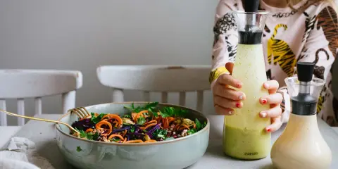 En skål med färgglad sallad och två flaskor dressing till sallad. En flicka håller i den ena flaskan.