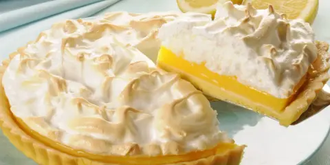 Puszysta tarta cytrynowa z bezą, z kawałkiem na łopatce. Obok leżą świeże cytryny.