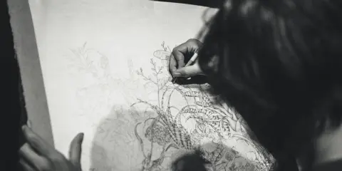 En konstnärs hand ritar med kol en detaljerad bild av växter och en insekt på ett stort papper.