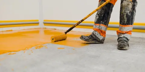 Un ouvrier en combinaison orange et grise, les bottes sales, utilise un rouleau pour peindre le sol d'un garage en jaune vif.