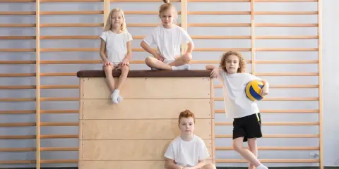 Dzieci w sali gimnastycznej. Czy fizjoterapeuta może wystawić zwolnienie z wf? Dwoje siedzi na skrzyni, jedno na niej, jedno na podłodze.