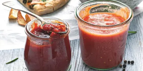 Dwa słoiczki domowego sosu do pizzy, jeden otwarty z łyżeczką, obok kawałek pizzy i przyprawy.