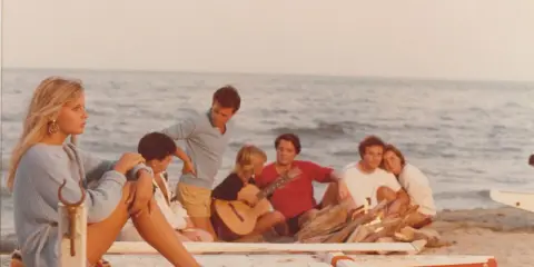 Giovani su una spiaggia, un gruppo di amici che ricorda i film estivi italiani, con chitarra e falò.