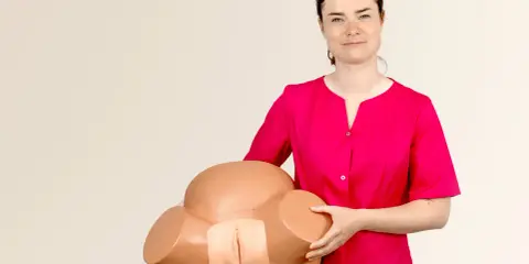 Kobieta w różowej bluzce trzyma model miednicy, demonstrując anatomię do nauki masażu szyjki macicy.
