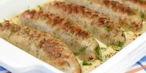 Biała kiełbasa zapiekana w sosie chrzanowym, udekorowana koperkiem, prezentuje się apetycznie w białym naczyniu.