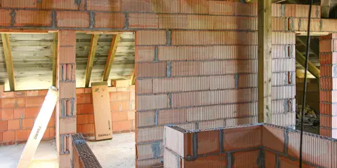 Niedokończone ściany z pustaków ceramicznych, widoczne drewniane belki stropowe. Wady murowania na piankę mogą prowadzić do problemów.