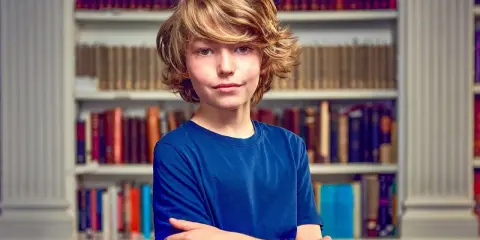 Garçon aux cheveux blonds, bras croisés devant une bibliothèque. Il semble prêt pour un test pour sauter une classe.