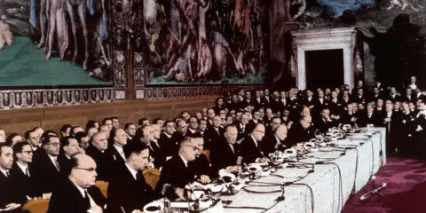 Uomini in abito scuro siedono a un lungo tavolo, con un murale storico sullo sfondo. Un momento storico, forse il **patto di Parigi**.