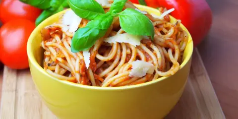 Pyszne spaghetti bez mięsa w żółtej misce, udekorowane bazylią i parmezanem. Obok pomidory i papryka.