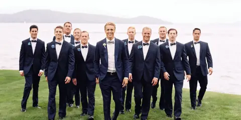 Groomsmen in navy suits with bow ties walk on grass by a lake. Can you wear a normal suit to a black tie event? This image shows a stylish alternative.