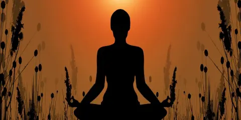 Silhuett av en person i meditationsställning, omgiven av gräs och med en solnedgång i bakgrunden. En bild som symboliserar meditation och hinduism.